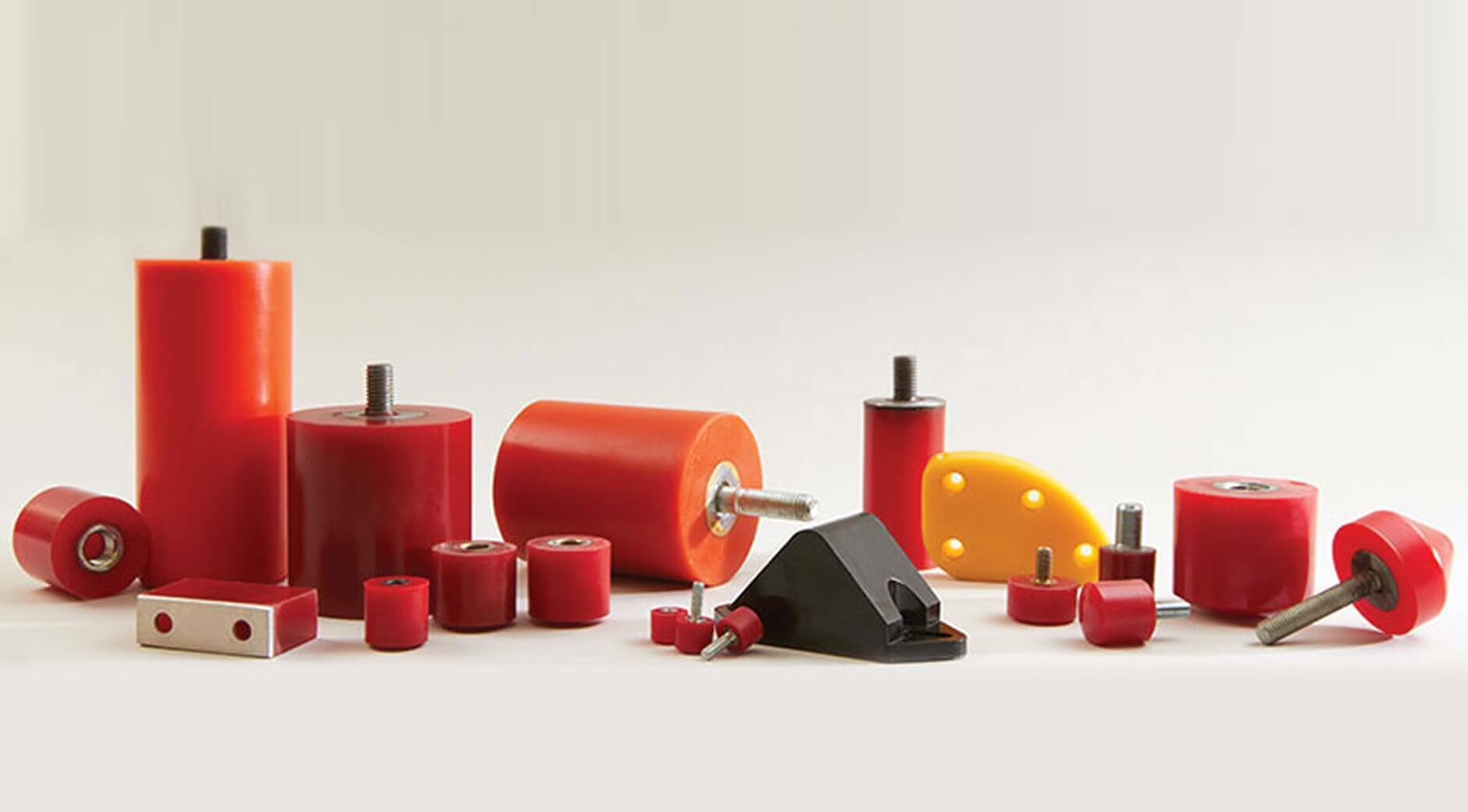 Assortment of polyurethane bumpers and mounts in various sizes and shapes, including cylindrical, rectangular, and curved styles, primarily in red and orange with metal threaded inserts and bolts, alongside a black metal bracket mount and a yellow urethane pad.